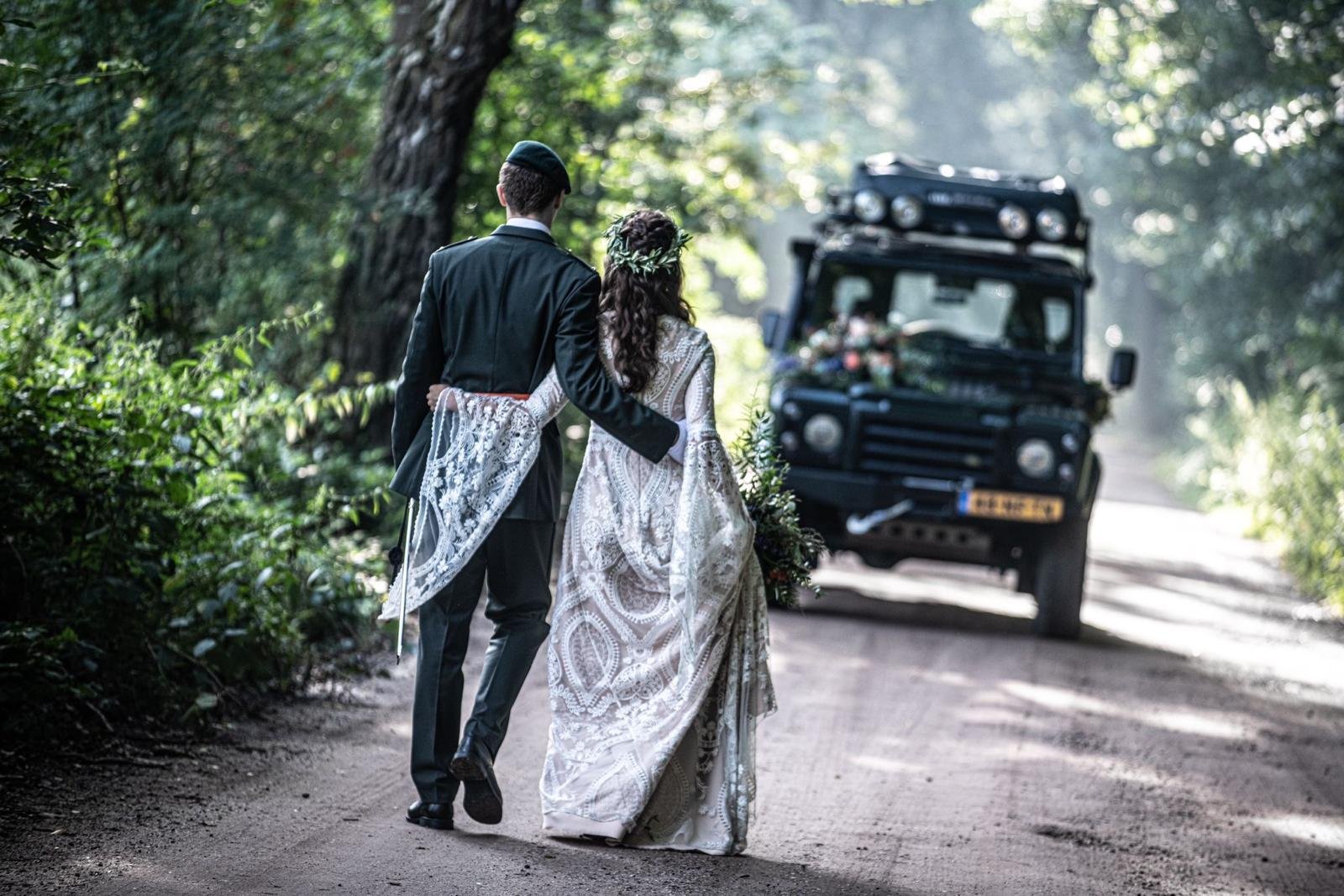 Bruidspaar loopt over een bospad naar de Land Rover Defender 110 als trouwauto